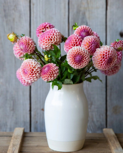 White vase with pink flowers on a wooden surface against a rustic wooden wall.