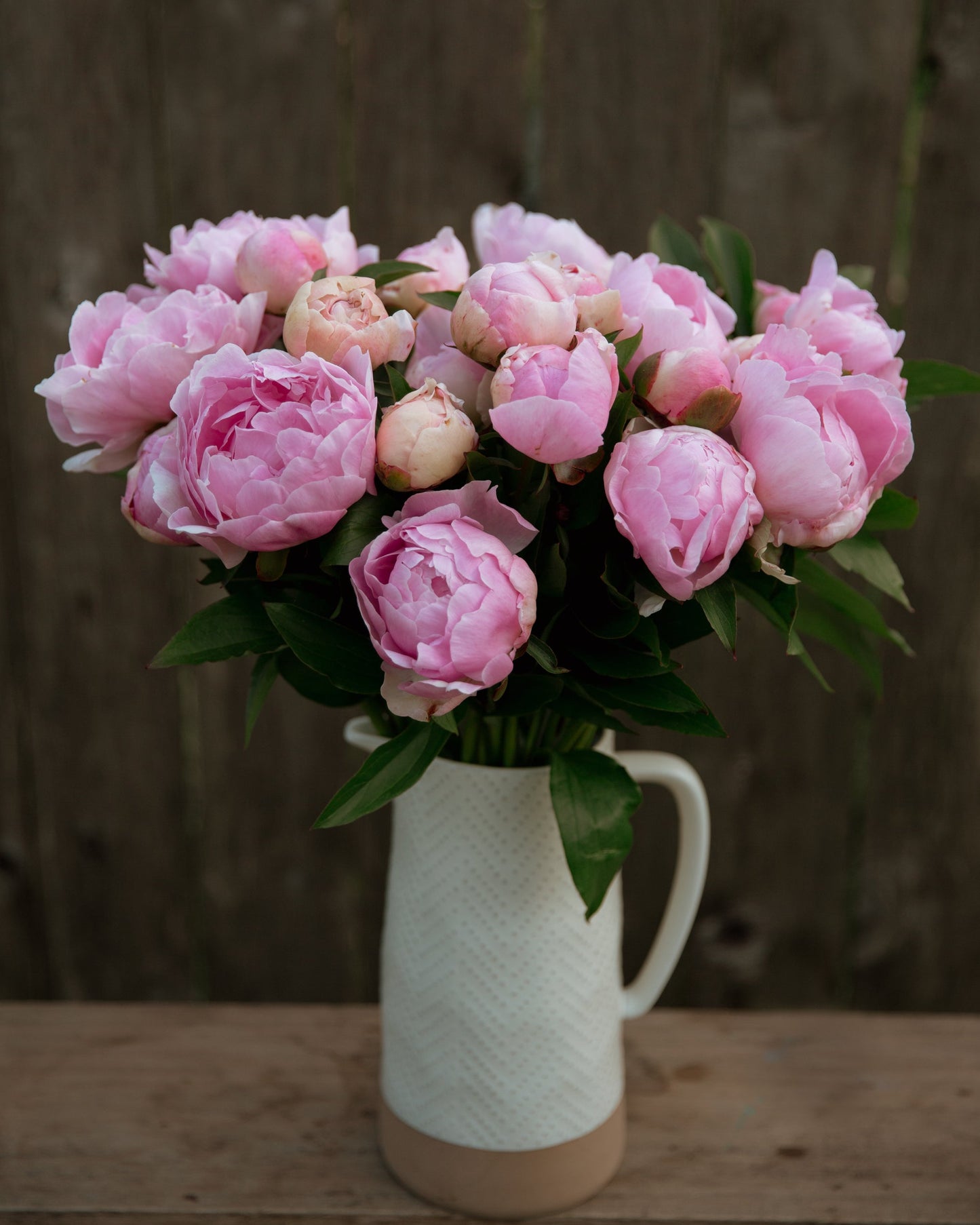 Bouquet of pink peonies in a white vase on a wooden surface with a wooden background