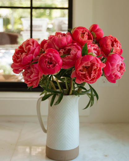 Bouquet of pink coral color peony flowers in a white vase on a marble surface with a window in the background.